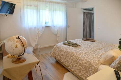 Immagine con la finestra della camera da letto al centro, una televisione e la scrivania a sinistra 